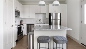 Une cuisine moderne avec des meubles blancs, des appareils en acier inoxydable, un comptoir en granit et deux tabourets de bar.
