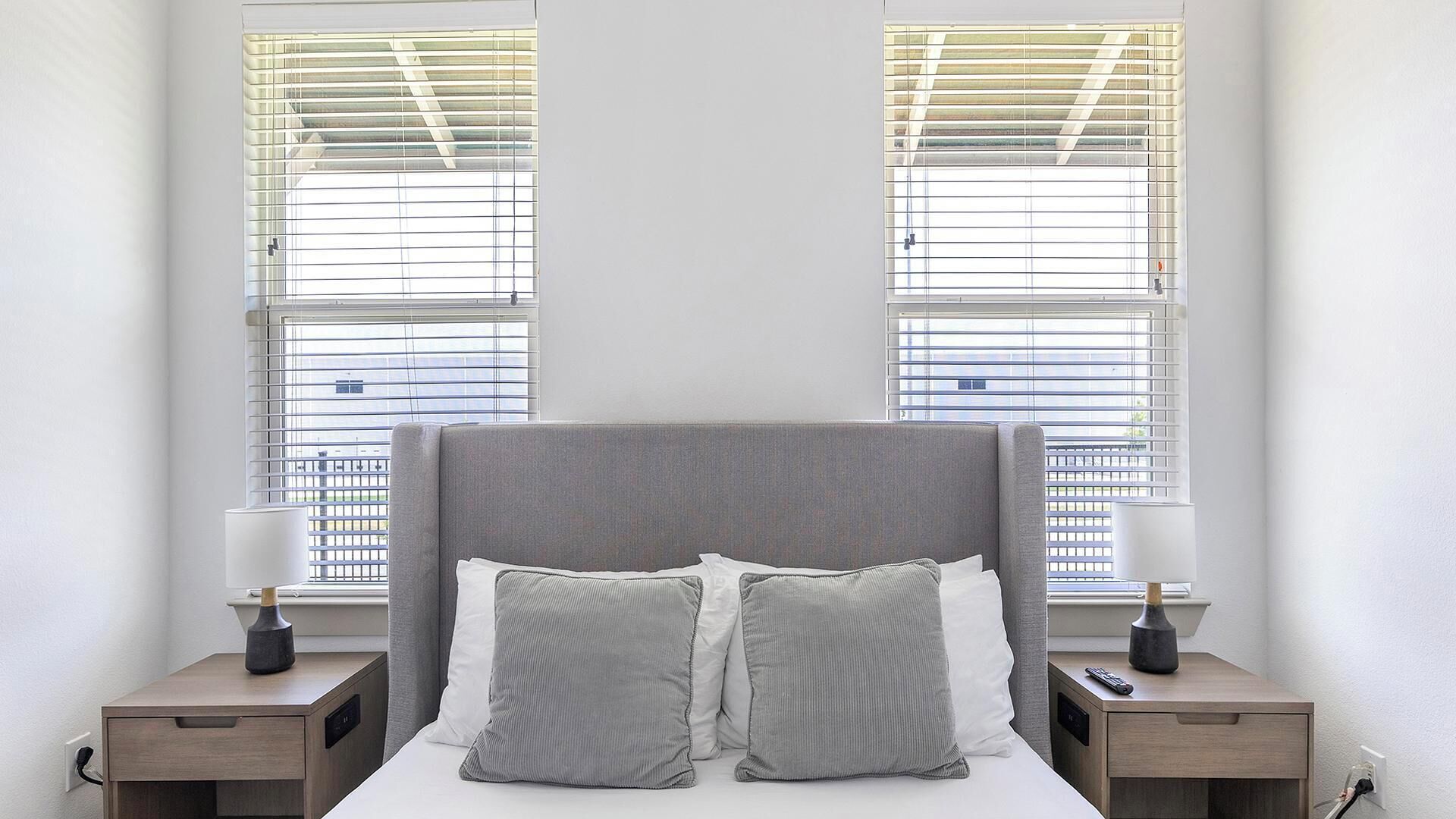 Une chambre avec une tête de lit grise, deux tables de chevet avec des lampes et deux fenêtres équipées de stores.