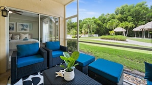 A covered outdoor seating area with wicker furniture and blue cushions, overlooking a well-maintained lawn and trees. - Limited-Time Offer: Charming 2BR Naples Condo with Club Option (Naples)