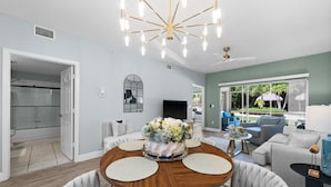 A modern living room with a round wooden table, white upholstered chairs, a chandelier, and a view of the outdoors. - Limited-Time Offer: Charming 2BR Naples Condo with Club Option (Naples)