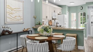 A dining area with a round table, white upholstered chairs, a chandelier, and a kitchen in the background. - Limited-Time Offer: Charming 2BR Naples Condo with Club Option (Naples)