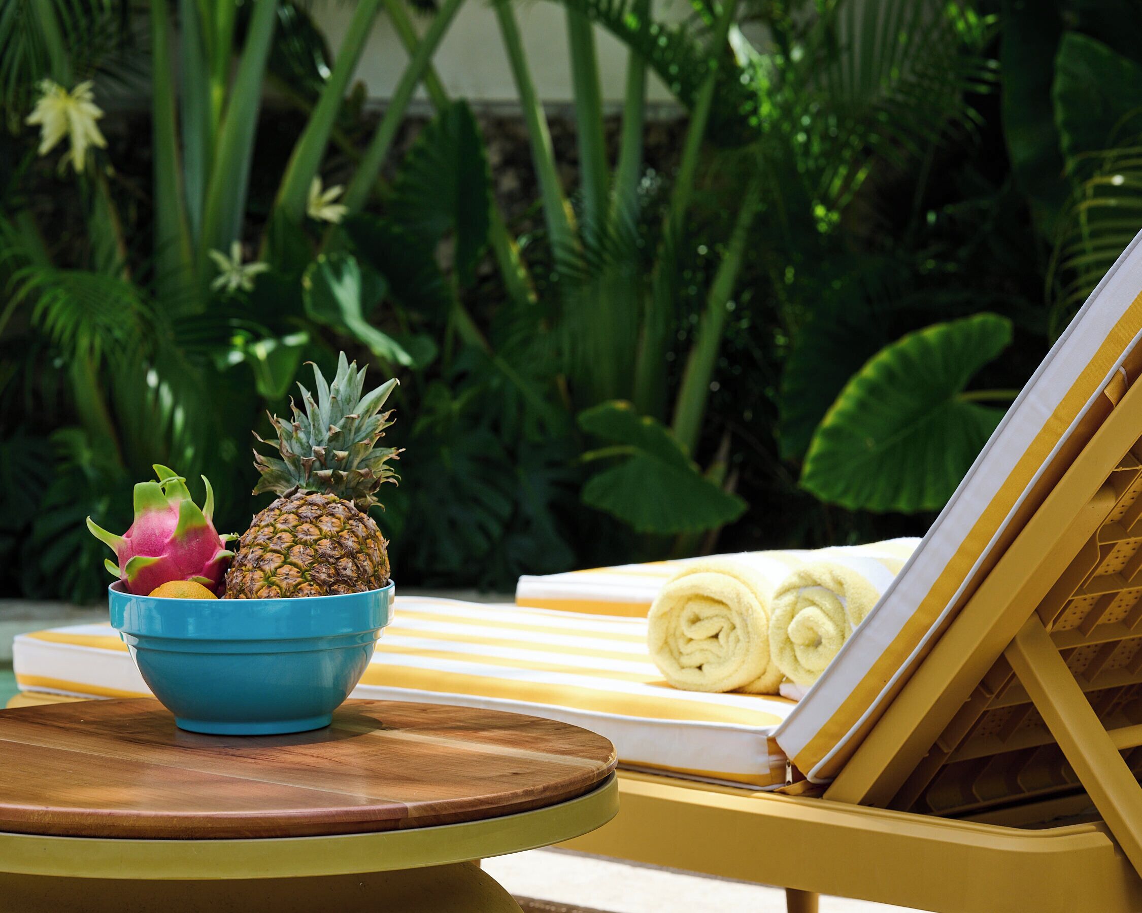 A tropical setting with a wooden table holding a bowl of fruit, a yellow lounge chair, and a rolled-up towel.