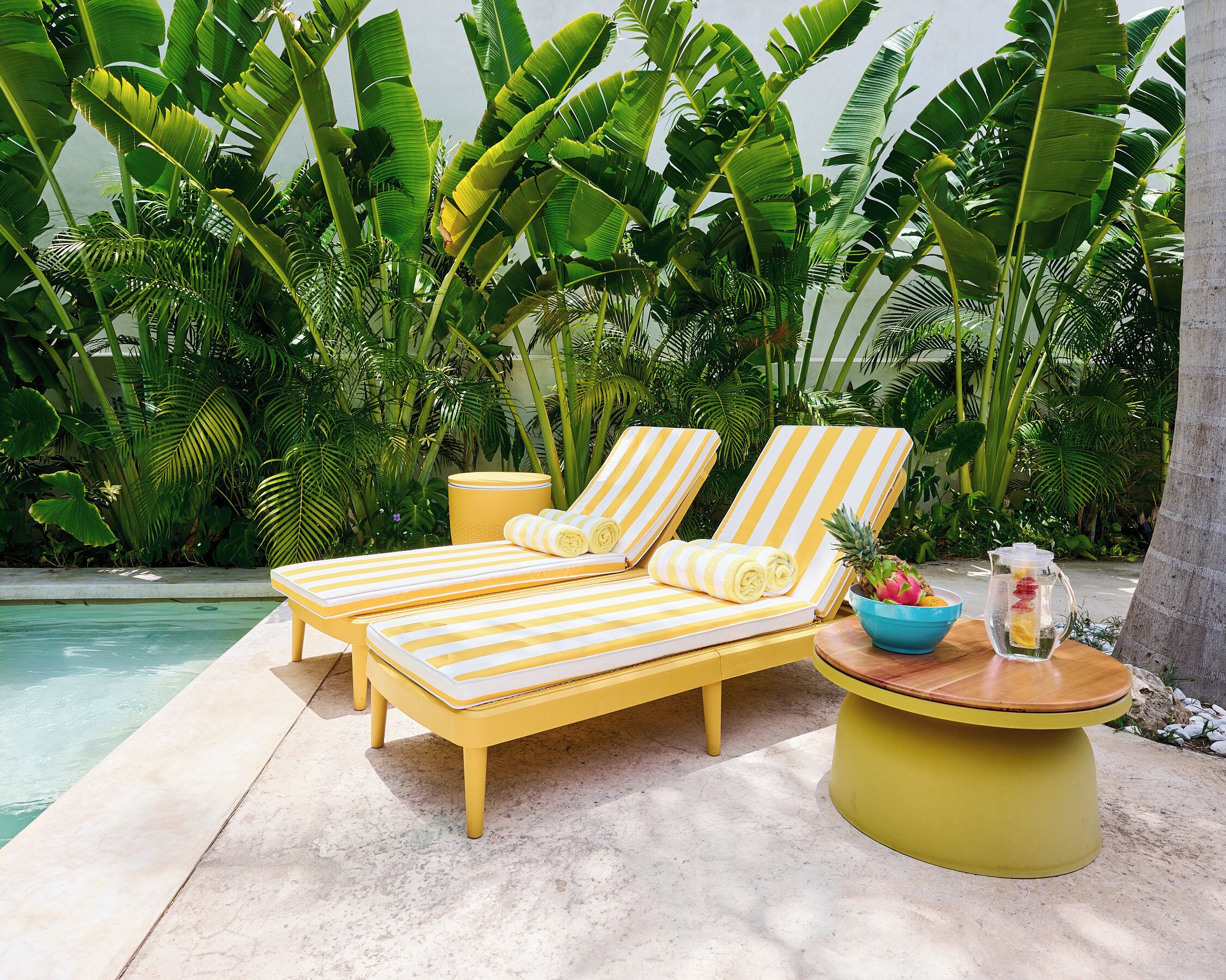 Un espace de détente aménagé au bord de la piscine, avec deux chaises longues à rayures jaunes, une petite table ronde jaune sur laquelle se trouvent un saladier et une carafe, ainsi qu'une petite poubelle jaune.