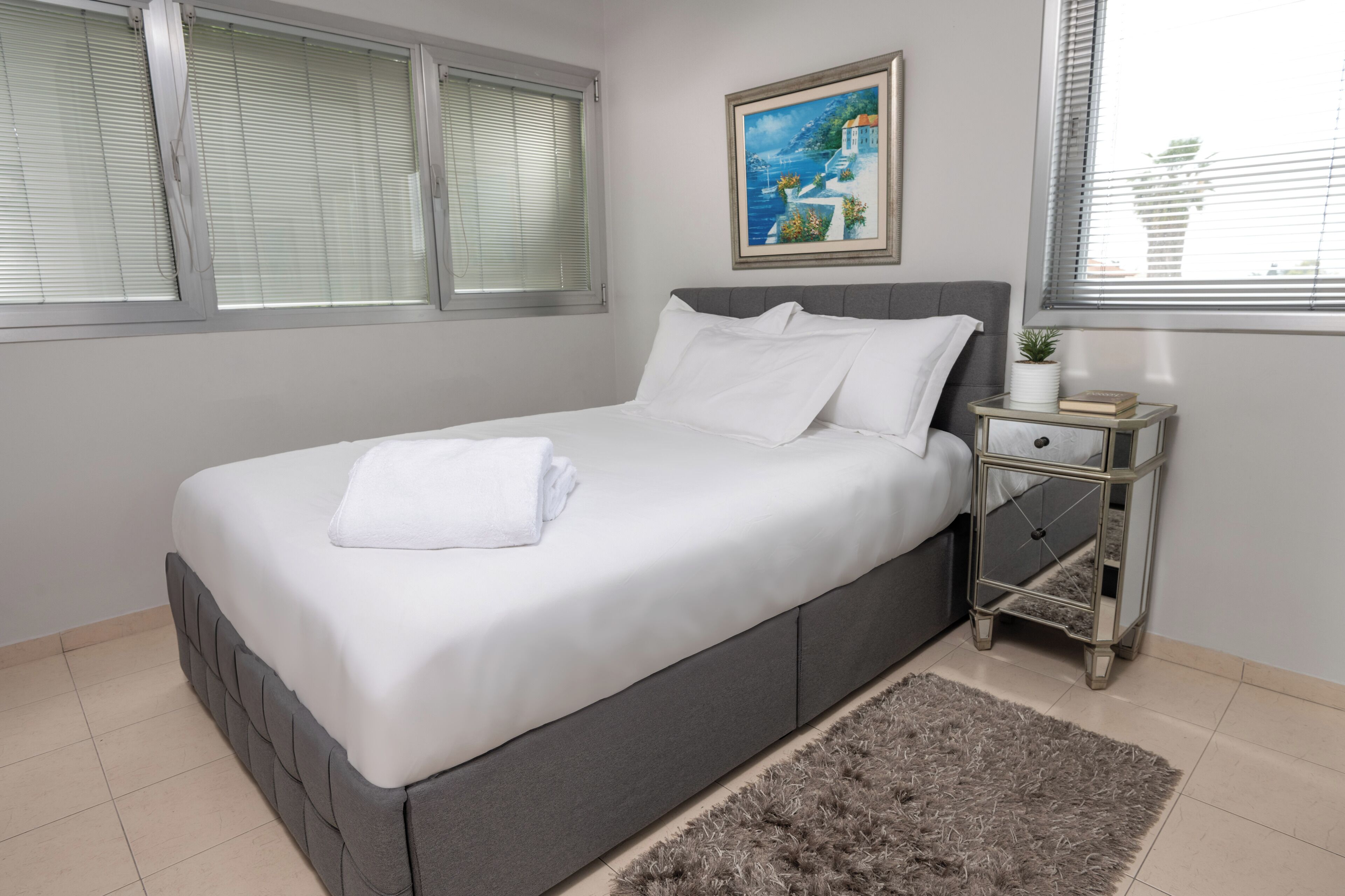 A bedroom with a bed, a nightstand, a painting, and a window with blinds.