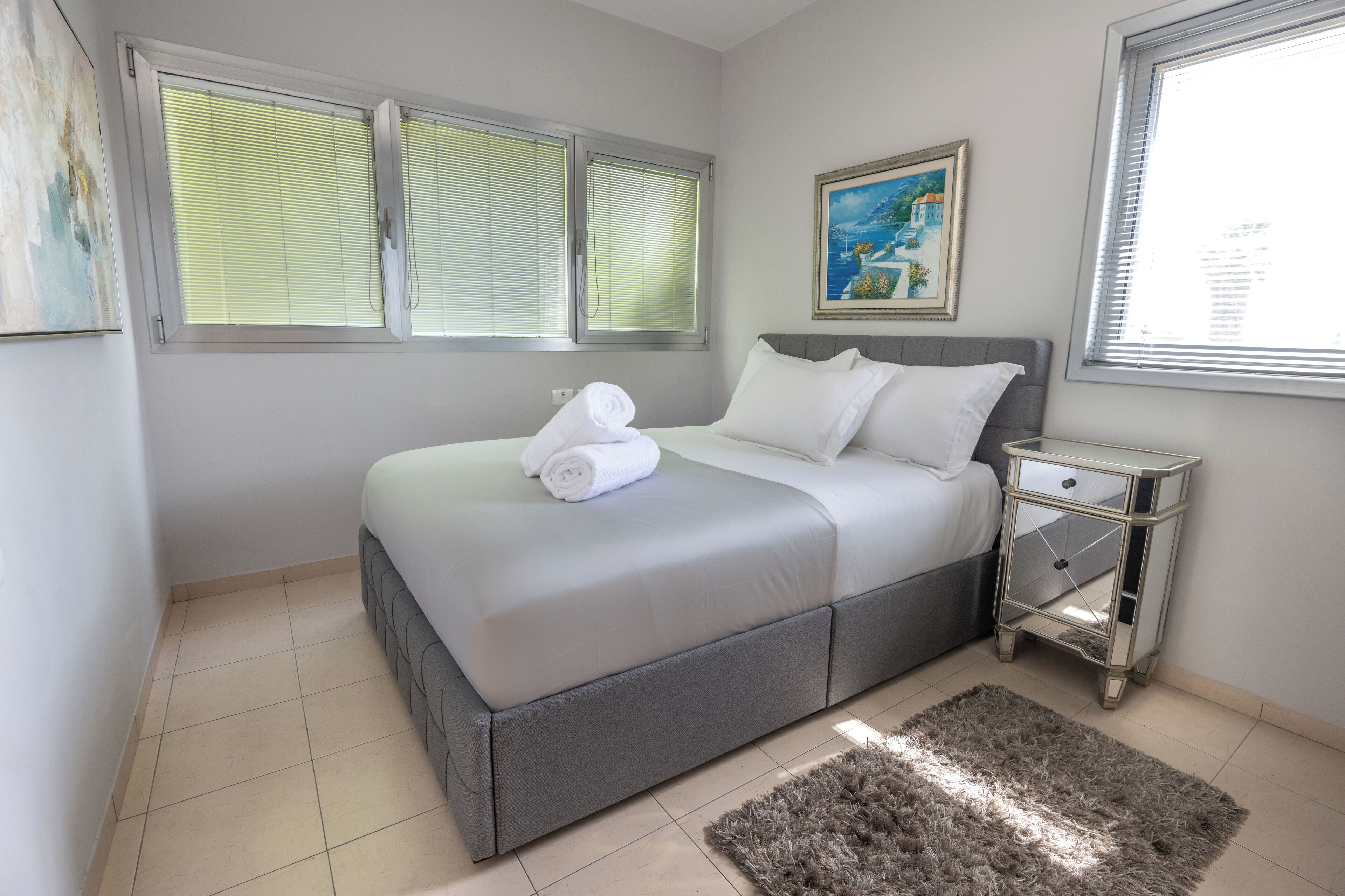 A bedroom with a bed, a nightstand, a painting, and a window with blinds.