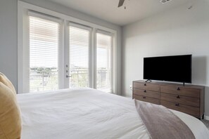 A bedroom with a large bed, a dresser with a flat-screen TV, and a balcony with white blinds.