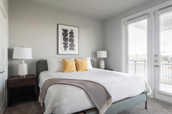 A neatly arranged bedroom with a bed, two bedside tables with lamps, a framed abstract artwork, and a view through the window.