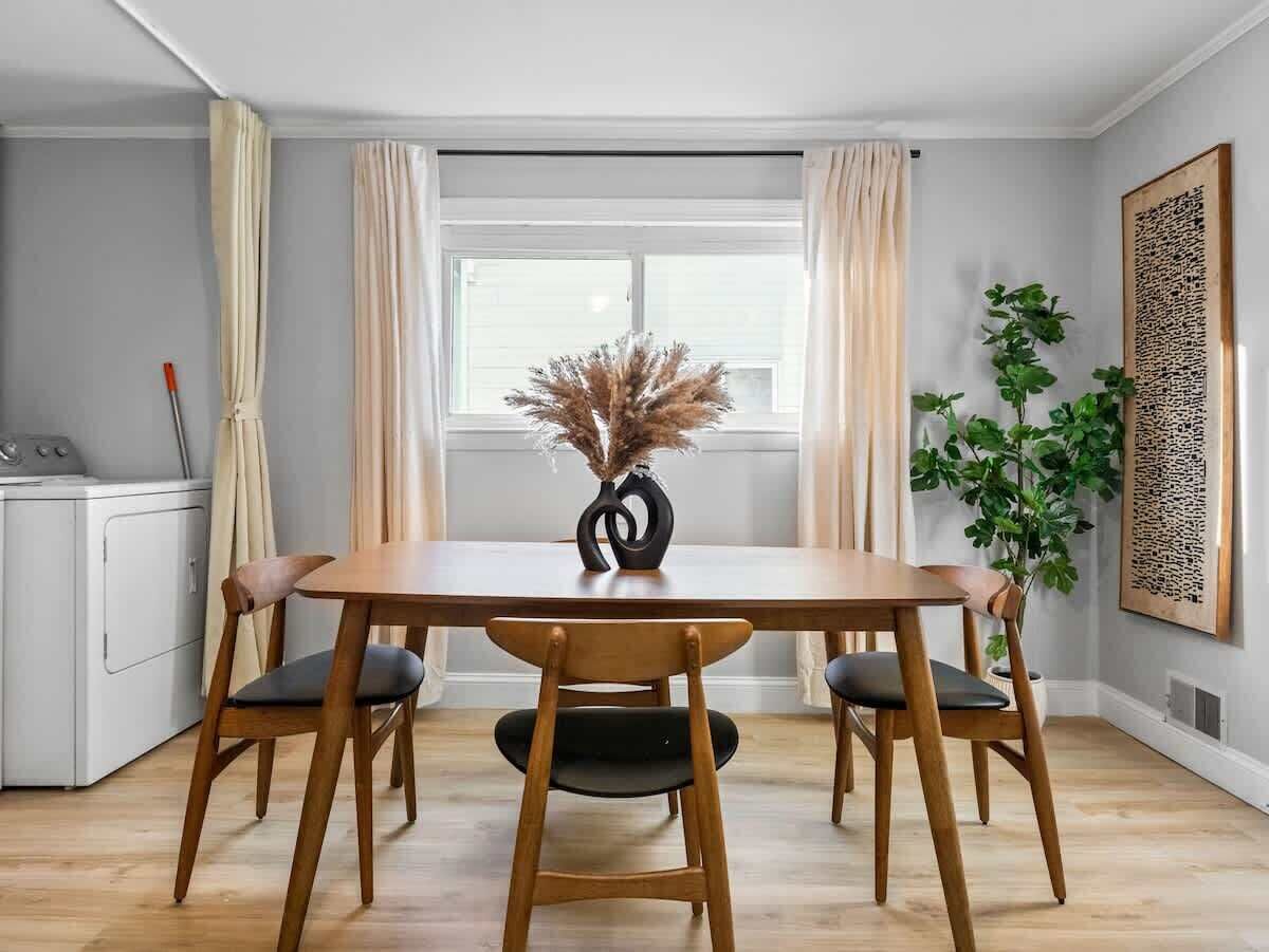 Een eetgedeelte met een houten tafel en stoelen, een vaas met gedroogde bloemen, een potplant en een kunstwerk aan de muur.