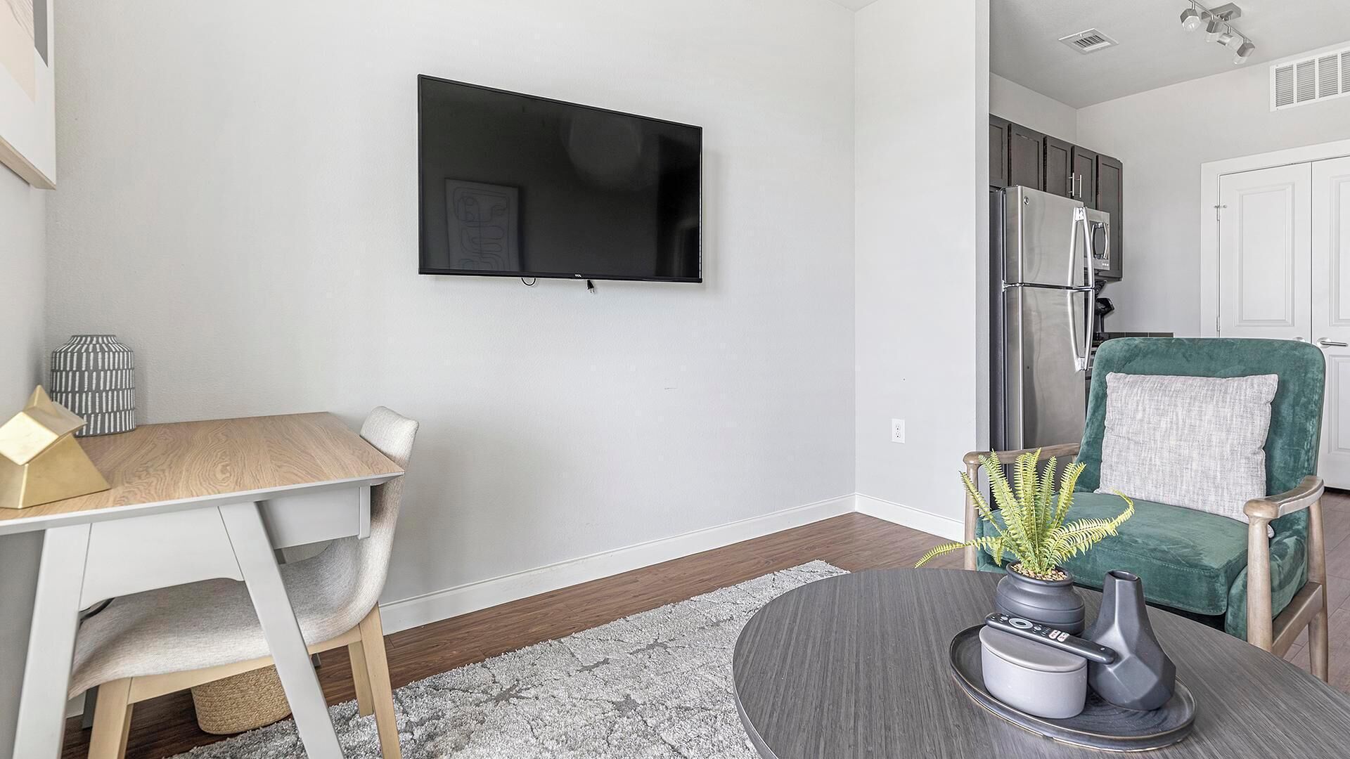 Een moderne woonkamer met een flatscreen televisie aan de muur, een houten eettafel met een stoel, een groene fauteuil met een plant, en een keuken met een koelkast en kasten.