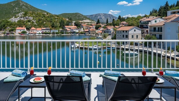 A balcony with outdoor seating, overlooking a lake with boats and a town in the background.