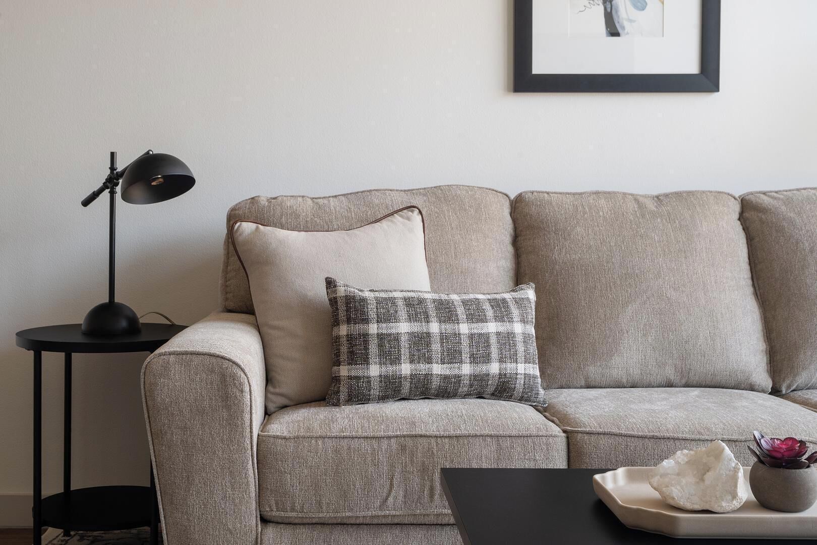 A grey sofa with a plaid pillow, a black floor lamp, and a small table with a vase and a rock.