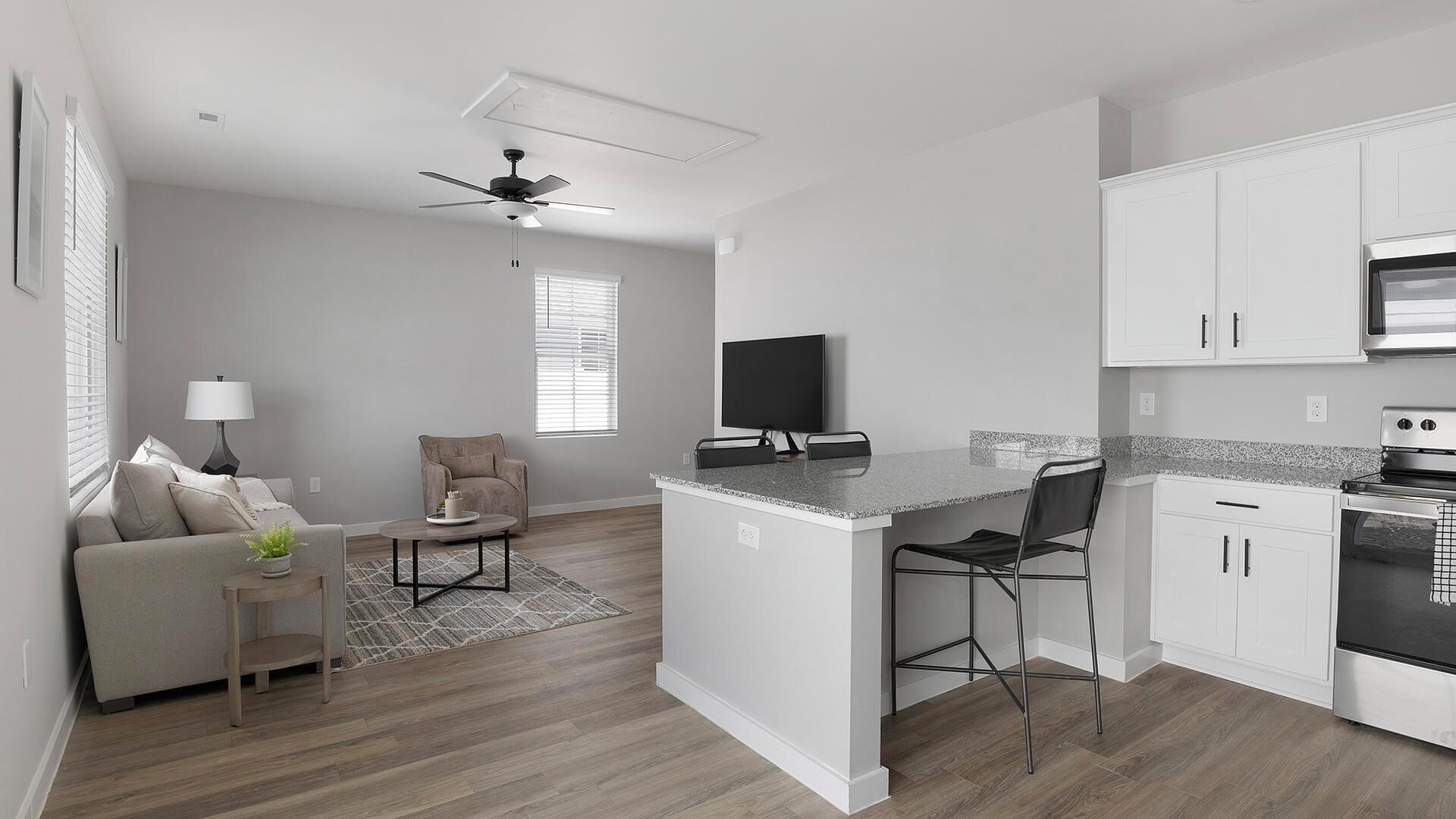 A modern kitchen with white cabinets, a built-in oven, and a microwave. There is a small island with bar stools and a flat-screen TV mounted above it.
