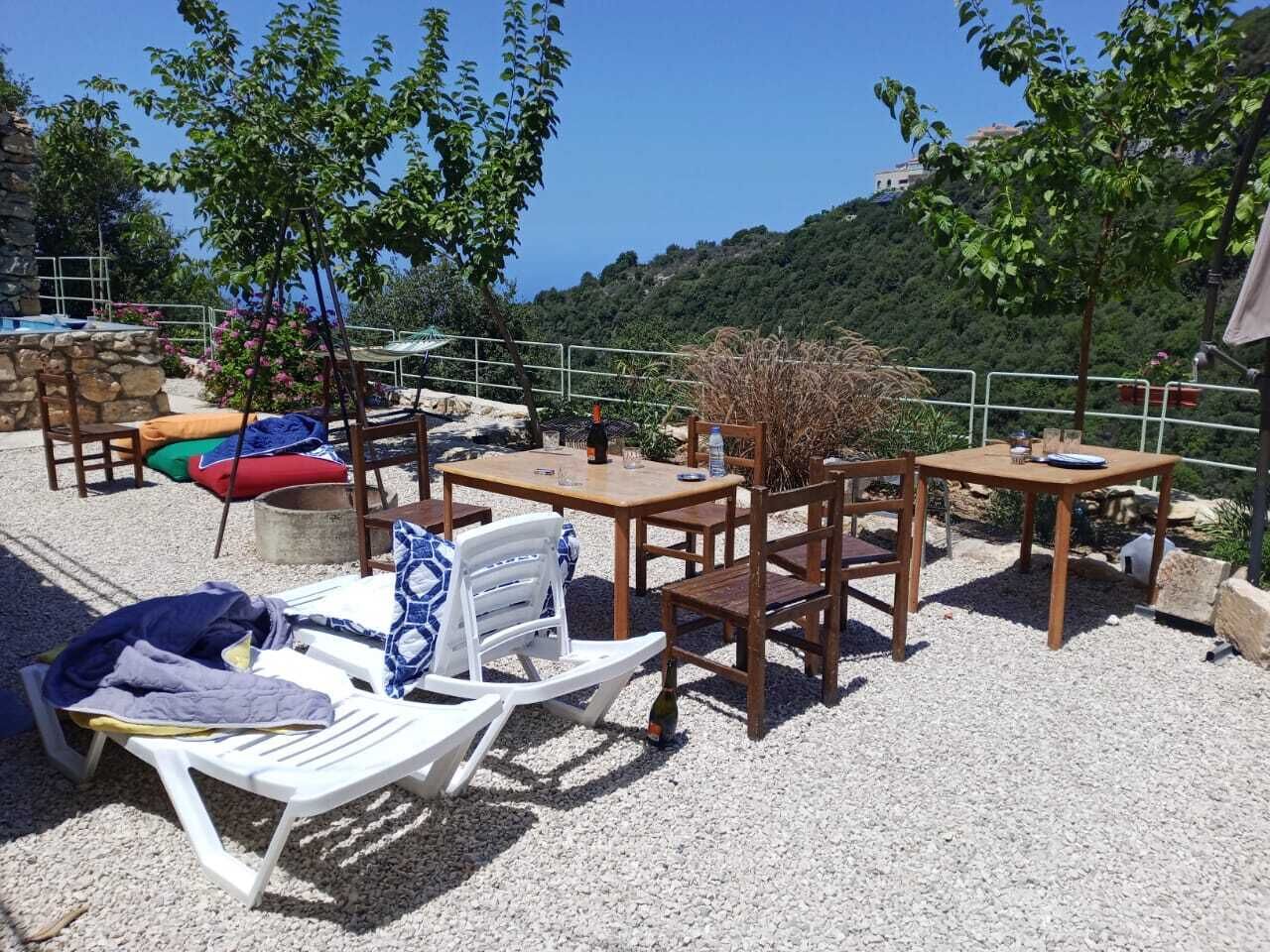 An outdoor seating area with tables and chairs, surrounded by gravel and greenery, overlooking a hillside.