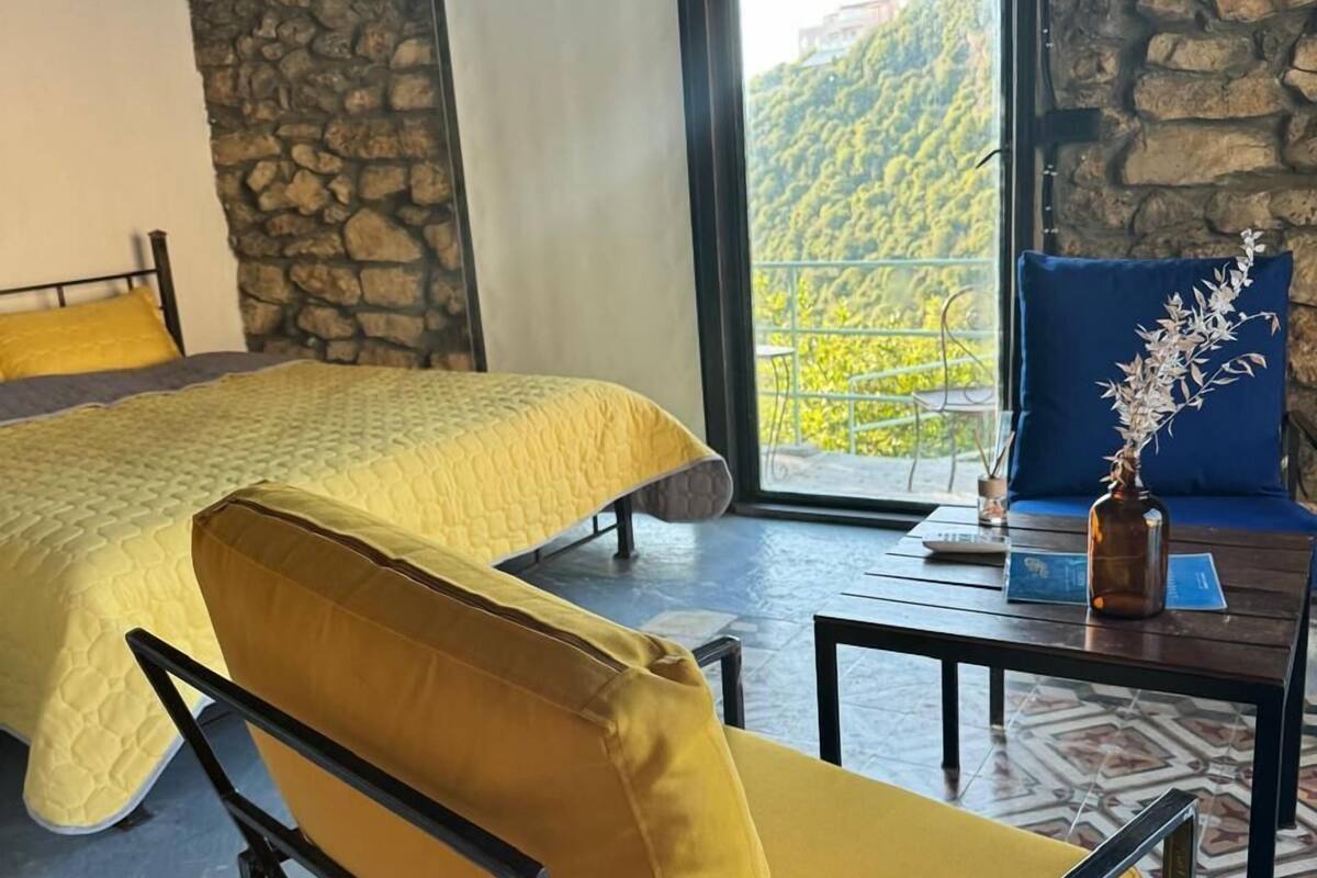 Una habitación con una cama, una silla amarilla, una mesa con un florero y vistas a la vegetación por una ventana.