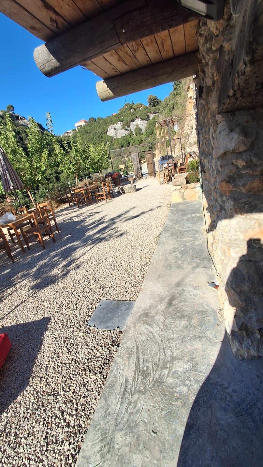 A rustic outdoor seating area with wooden tables and chairs, a gravel path, and a stone wall.