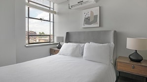 Un dormitorio con cama, mesita de noche, lámpara, ventana con persianas y aire acondicionado instalado en la pared.