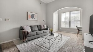 A modern living room with a grey sofa, a glass coffee table, and a large window with blinds.
