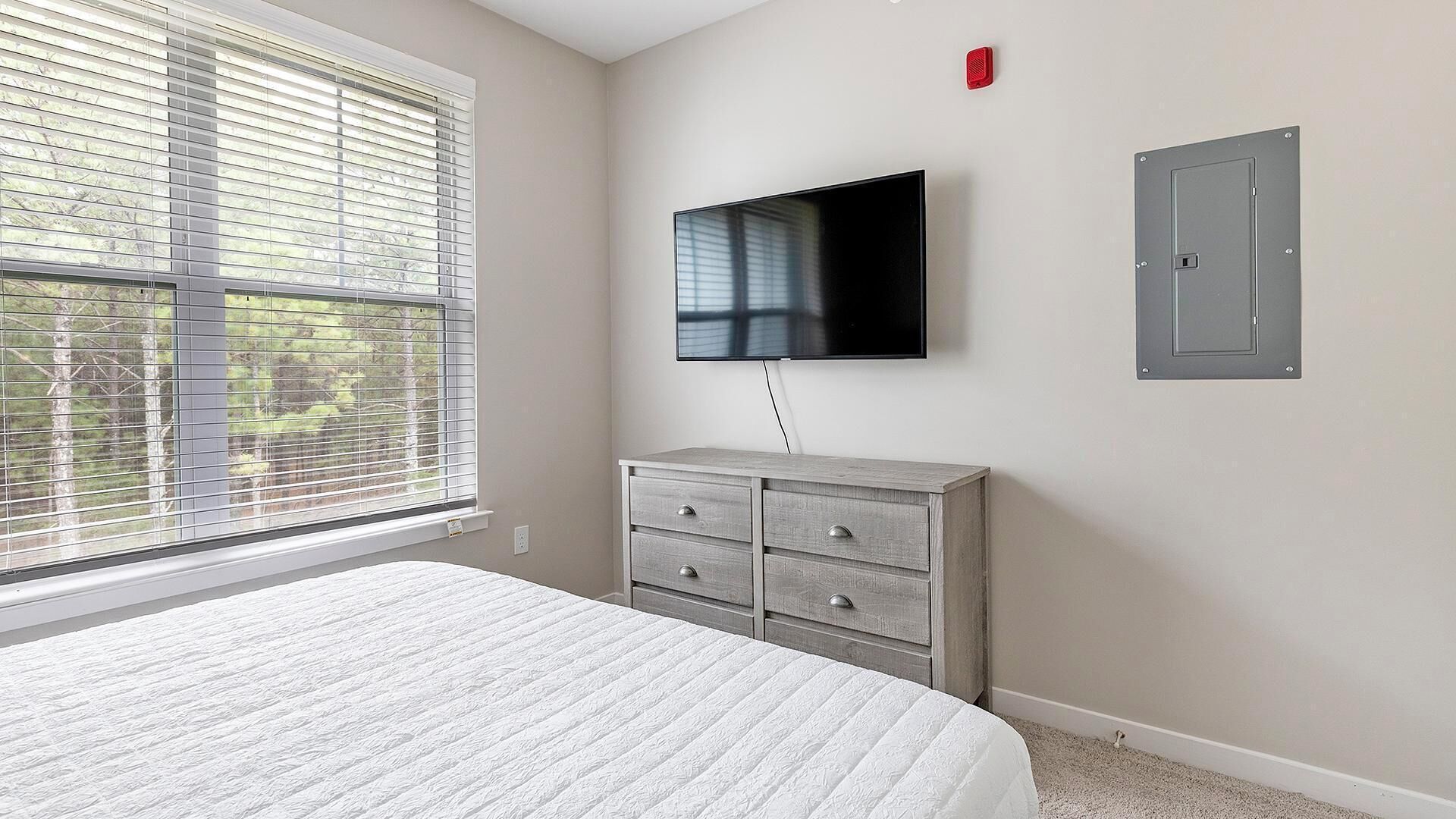 Une chambre à coucher comprenant un lit, une commode, un téléviseur fixé au mur et une fenêtre avec des stores.
