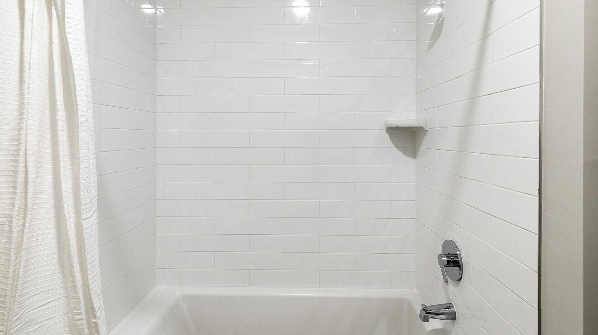 Une cabine de douche aux murs recouverts de carreaux blancs, avec une baignoire, un porte-serviettes d'angle et un rideau de douche.