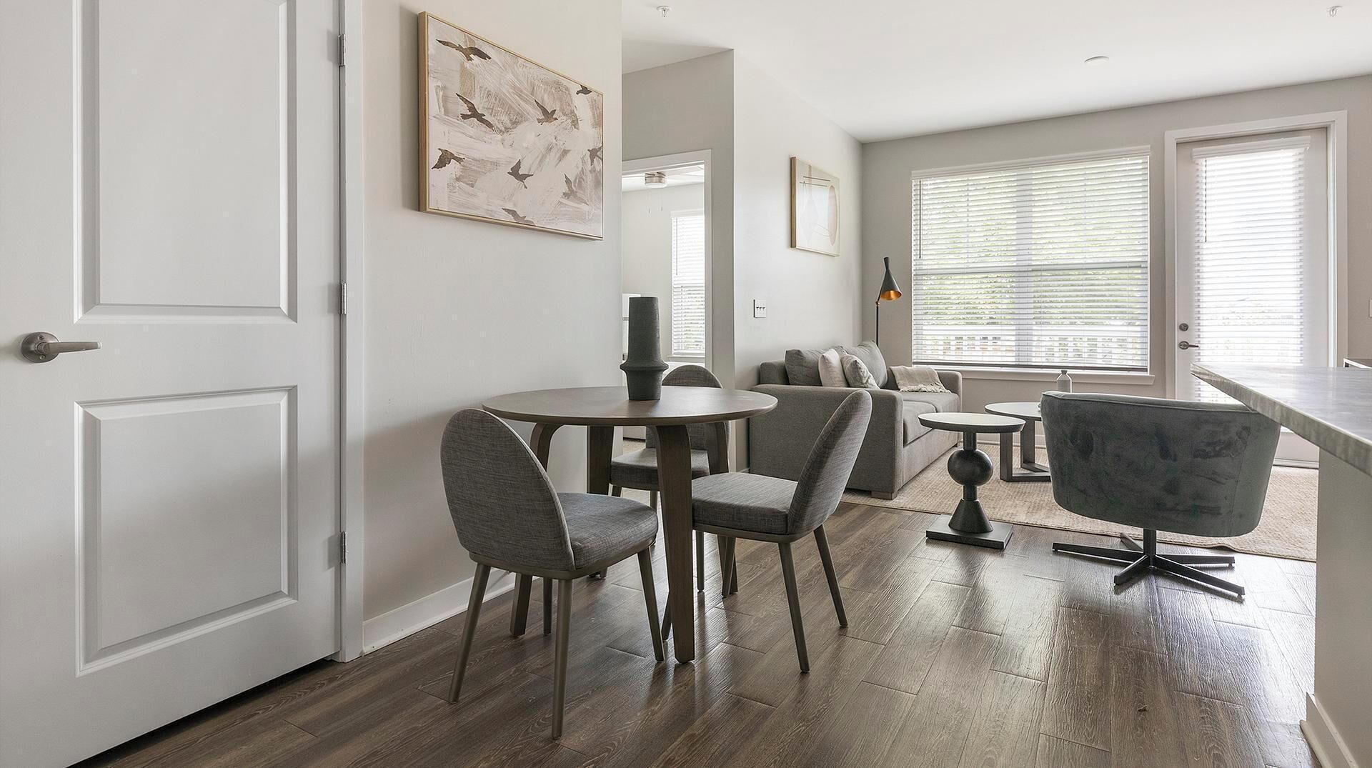 A modern living room with a sofa, a dining table with chairs, and a large window with blinds.