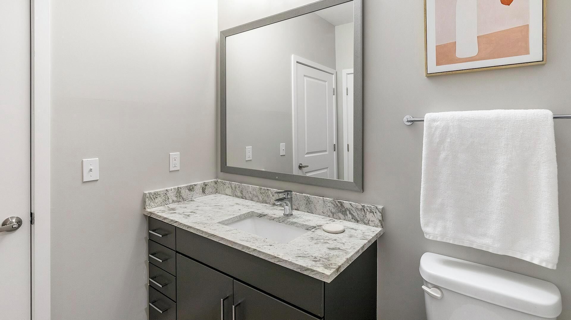 A bathroom with a marble sink, dark cabinets, a white toilet, and a framed picture on the wall.