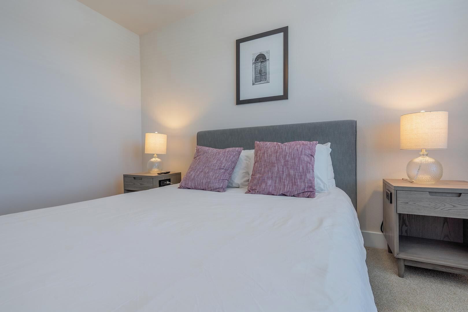 A neatly made bed with white linens and two purple pillows, flanked by two bedside tables with lamps, one of which has a decorative glass lampshade.