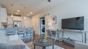 A modern living room with a sofa, a coffee table, and a TV. There is a kitchen area with cabinets and a washing machine. - Landing | Amazing 1BD, Yoga Studio, Gym (San Antonio)