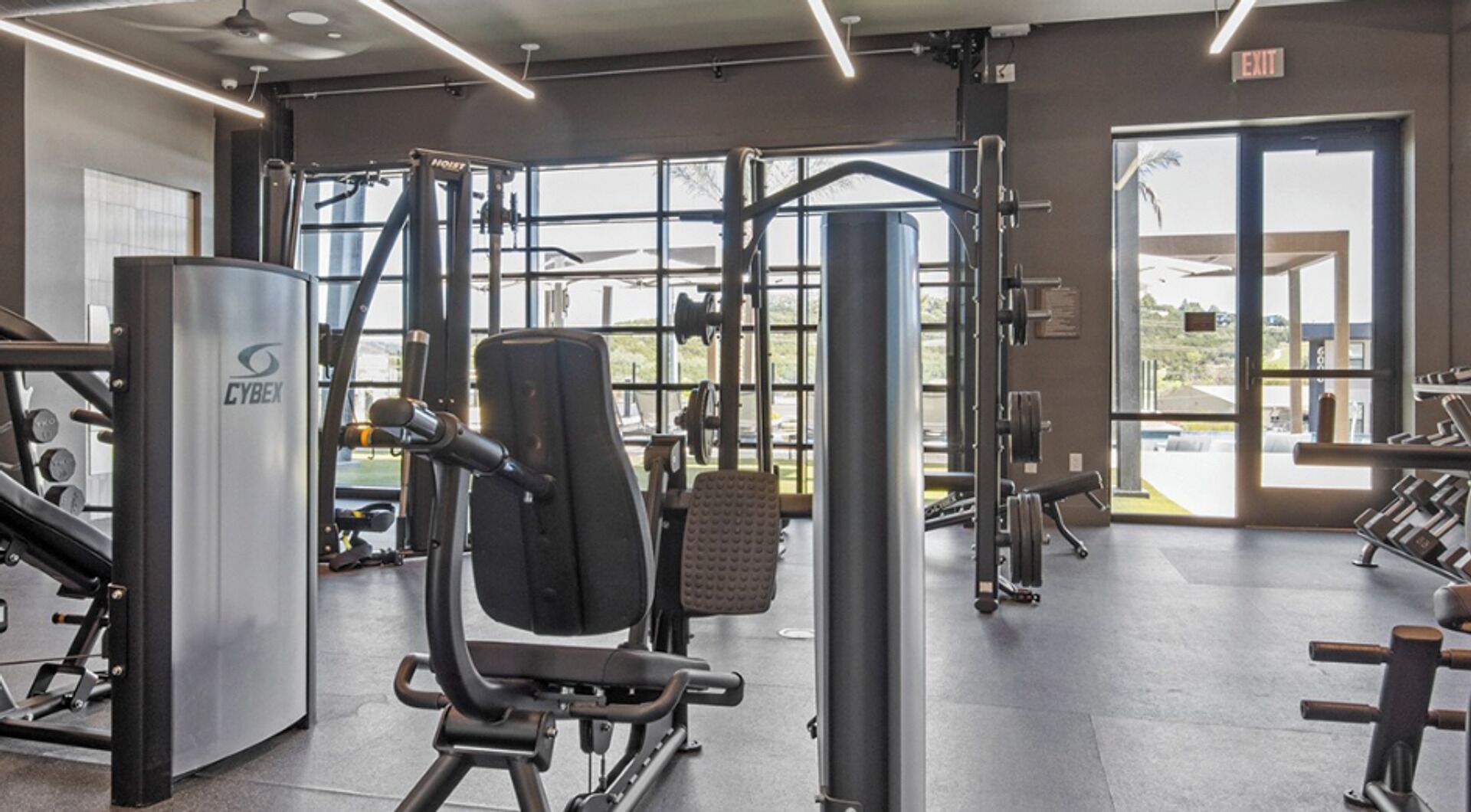 Ein Fitnessstudio mit verschiedenen Trainingsgeräten, großen Fenstern und einem Ausgangsschild.
