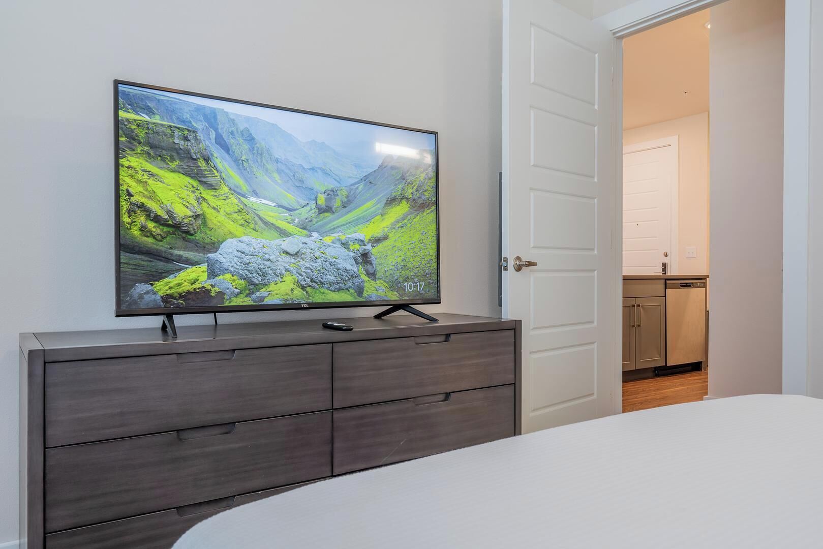 Une chambre à coucher avec un grand téléviseur à écran plat posé sur une commode en bois foncé, un lit blanc et une porte blanche donnant sur un coin cuisine.