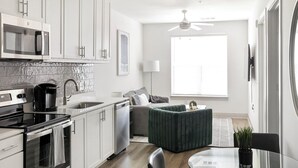 Una cocina moderna con armarios blancos, un microondas, una cafetera y una estufa. En la sala de estar hay un sofá gris y un reposapiés verde. También se puede ver un ventilador de techo y una ventana con persianas.