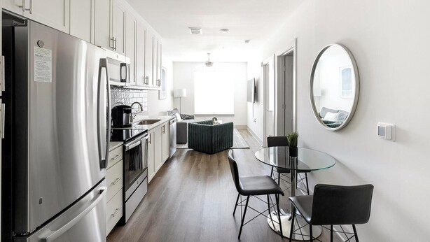 Una cocina moderna con electrodomésticos de acero inoxidable, una zona de comedor con mesa y sillas de vidrio, y un espejo grande en la pared.