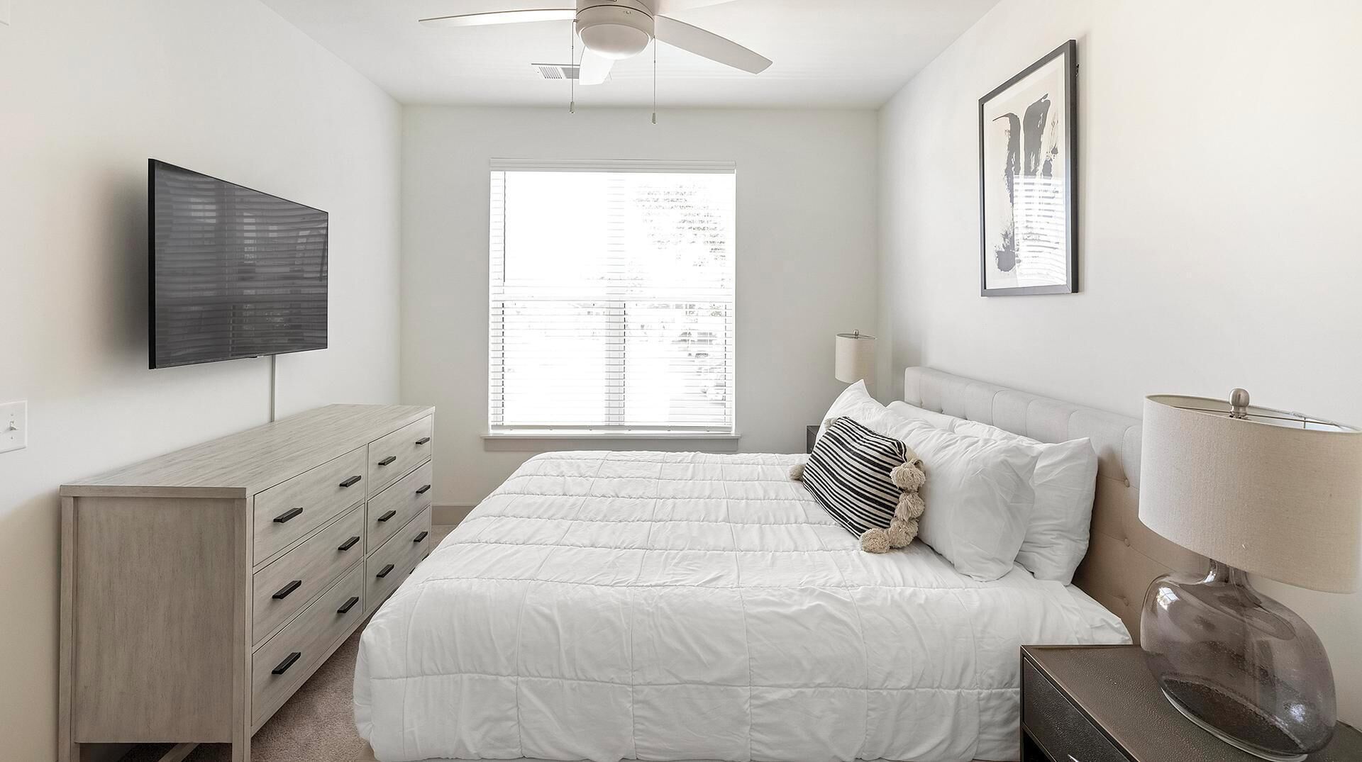 Un dormitorio con cama, cómoda, ventilador de techo, ventana y televisor instalado en la pared.