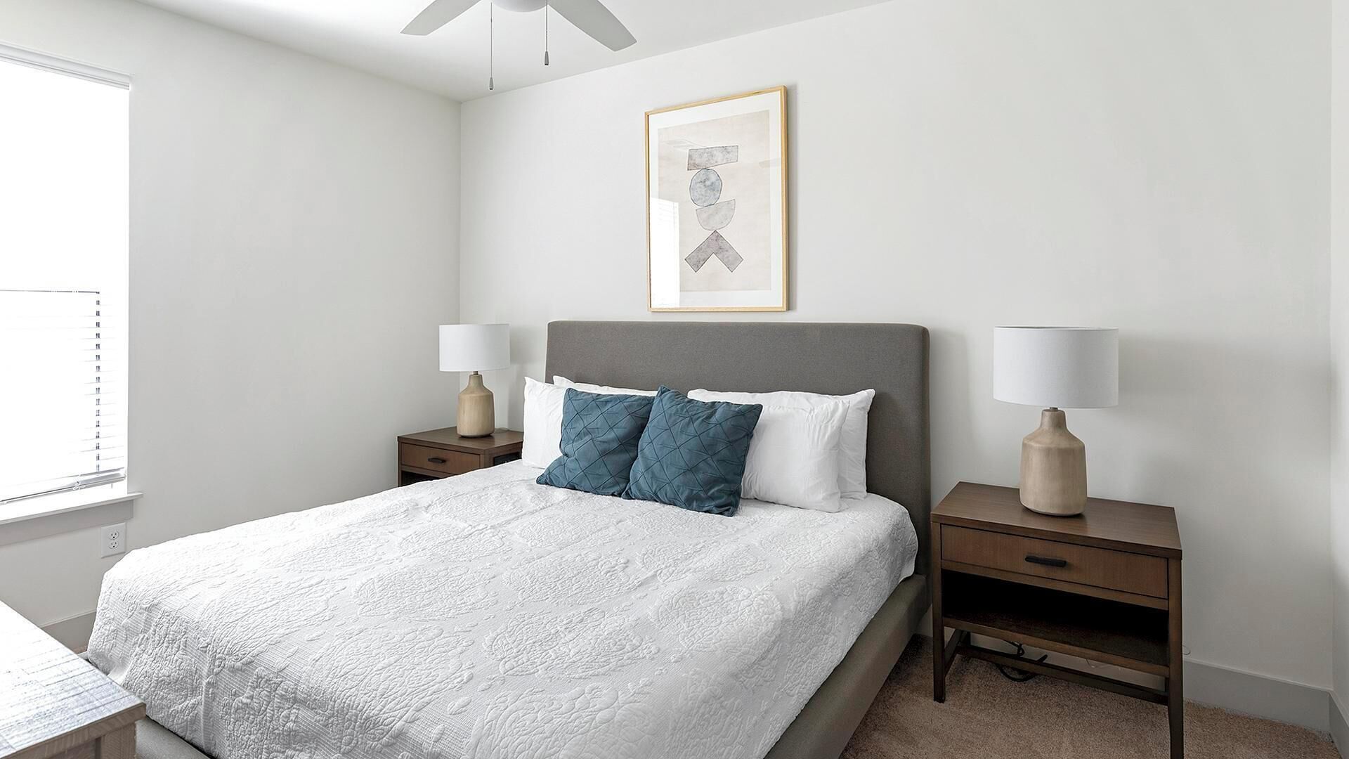 Un dormitorio ordenado con una cama grande, dos mesitas de noche con lámparas, una obra de arte enmarcada sobre la cama y una ventana con persianas.