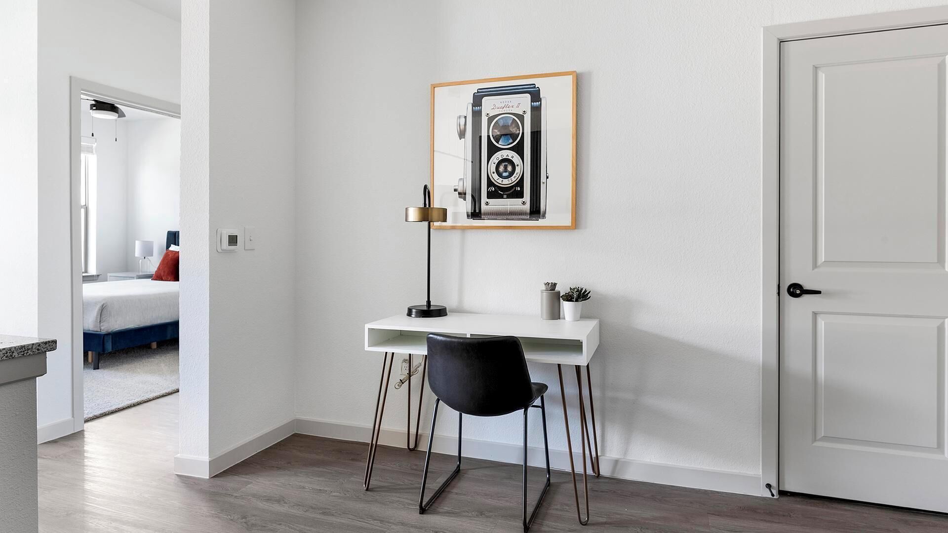 Una habitación con un escritorio blanco, una silla negra, una cámara antigua enmarcada en la pared y una puerta cerrada.
