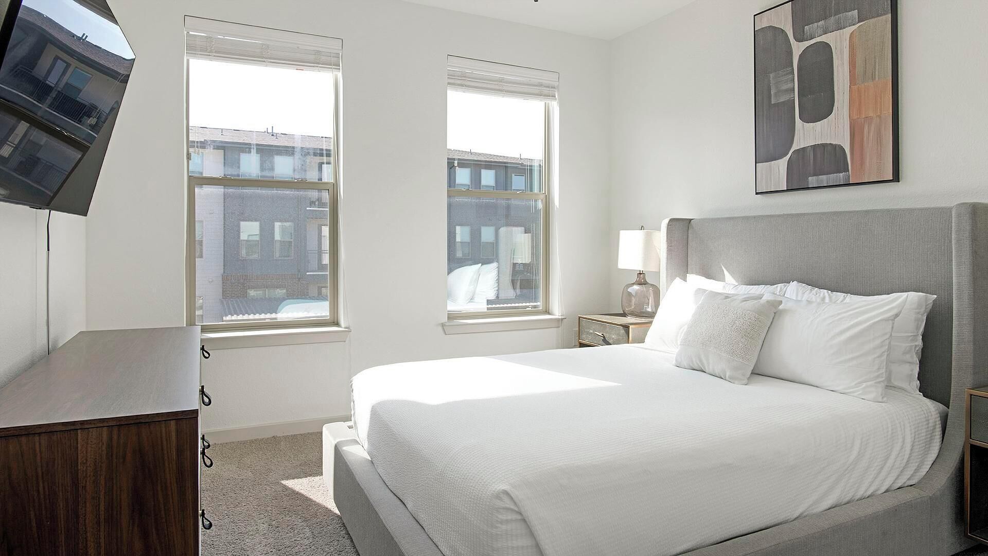 A hotel room with a large bed, two windows, a flat-screen TV, and a painting on the wall.