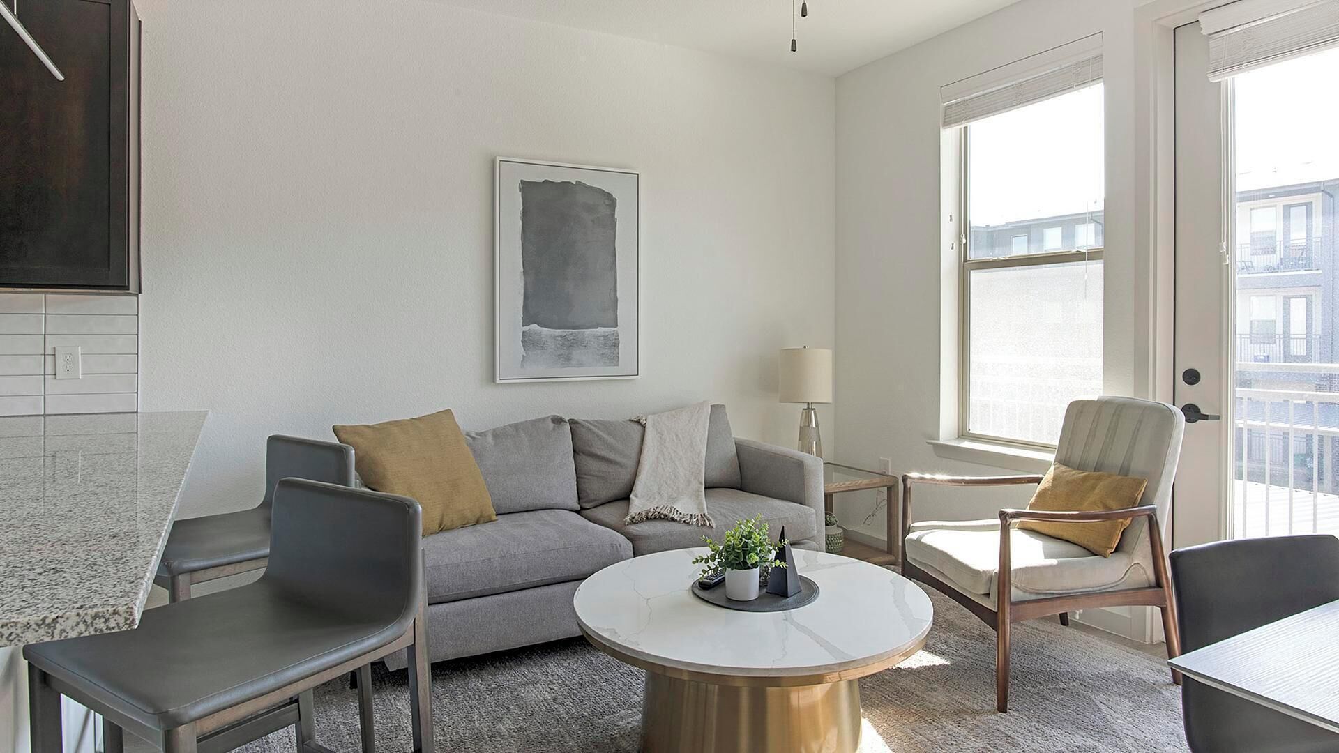 A modern living room with a grey sofa, two armchairs, a round coffee table with a plant, and a large window providing a view of buildings.
