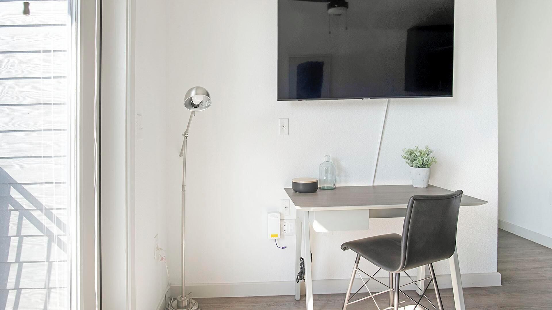 Une chambre moderne avec un mur blanc, un téléviseur à écran plat fixé au mur, un bureau avec une chaise, une lampe sur pied et une plante en pot.