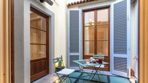 A small patio area with a glass table and chairs, a cactus, and a window with shutters.