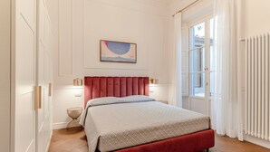 A bedroom with a bed, a nightstand, a lamp, a painting, and a window with curtains.