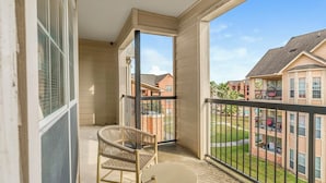 A balcony with a wicker chair and a small table, overlooking a residential area with brick buildings and a lawn.