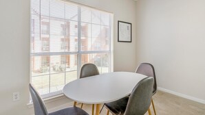Een eethoek met een ronde tafel en vier stoelen, grote ramen met luxaflex en een lijst met een foto aan de muur.