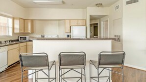 A modern kitchen with wooden cabinets, a white refrigerator, a microwave, and a granite countertop. There are three metal bar stools in front of the counter. - Landing | Chic 2BD, Clubhouse, Game Room (Pasadena)