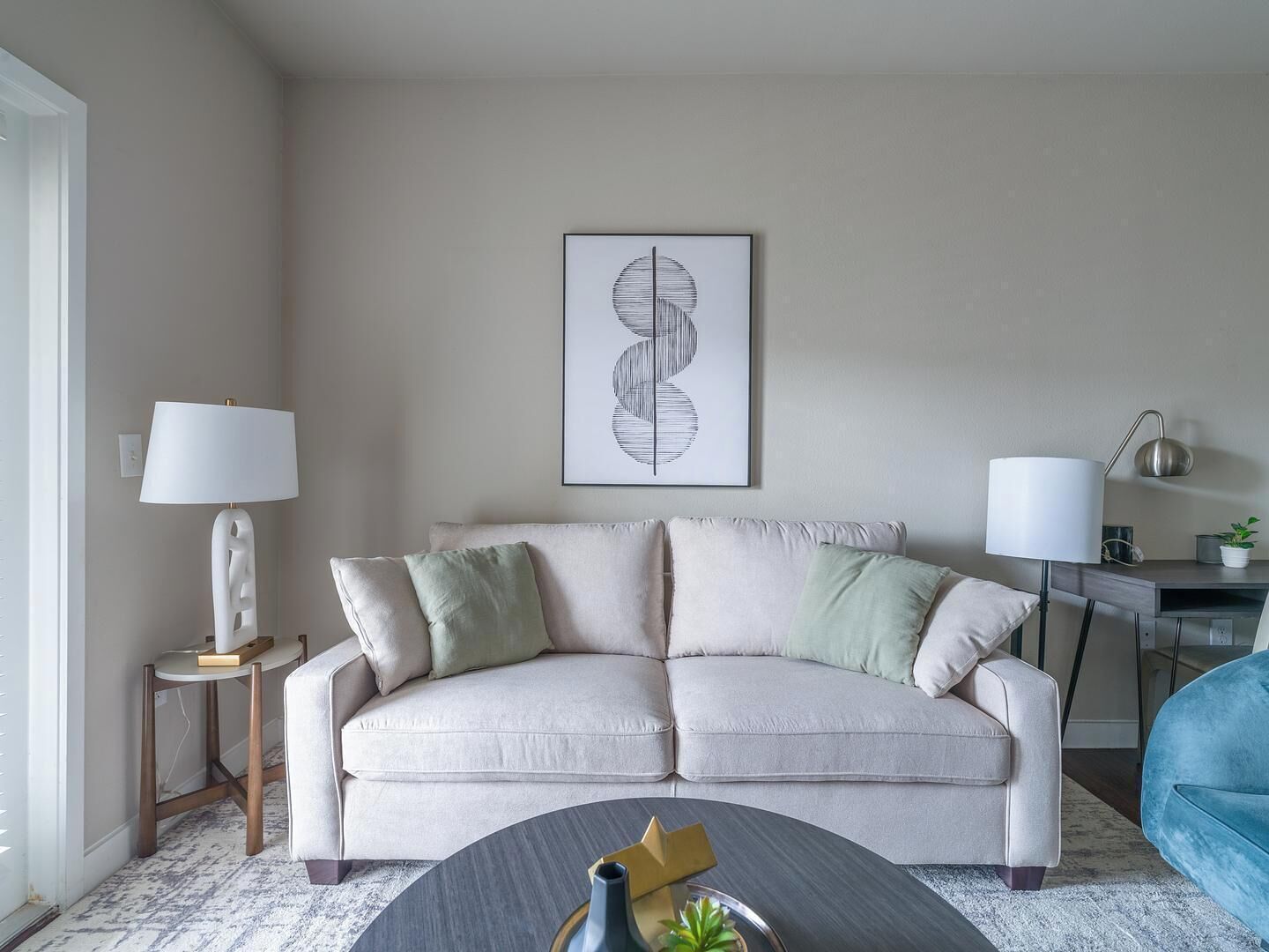 Un salon avec un canapé beige, deux tables d’appoint avec des lampes, une table basse ornée d’un vase et d’une plante, et une œuvre d’art abstraite encadrée accrochée au mur.
