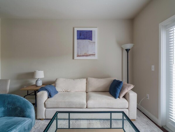 A living room with a beige sofa, a blue armchair, a glass coffee table, a floor lamp, and a framed artwork on the wall.
