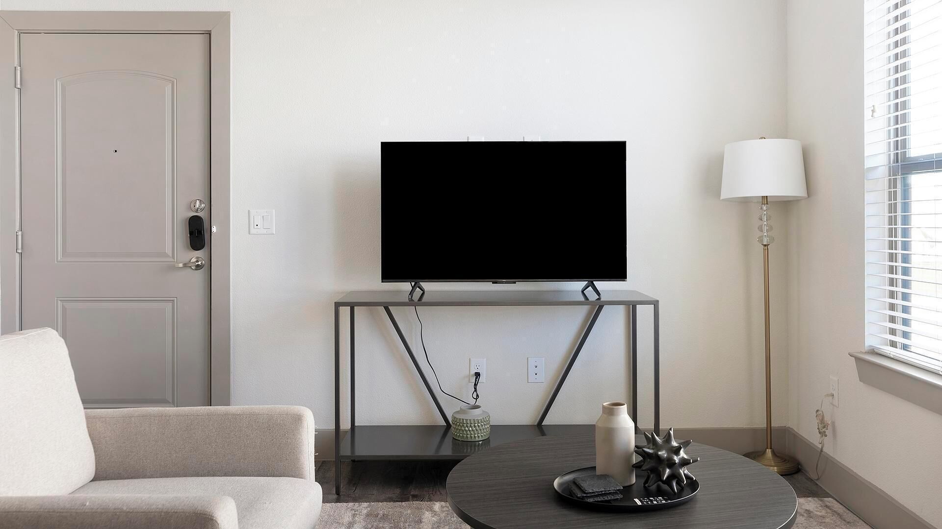 A modern living room with a flat-screen TV on a stand, a sofa, a coffee table with decorative items, a lamp, and a door.