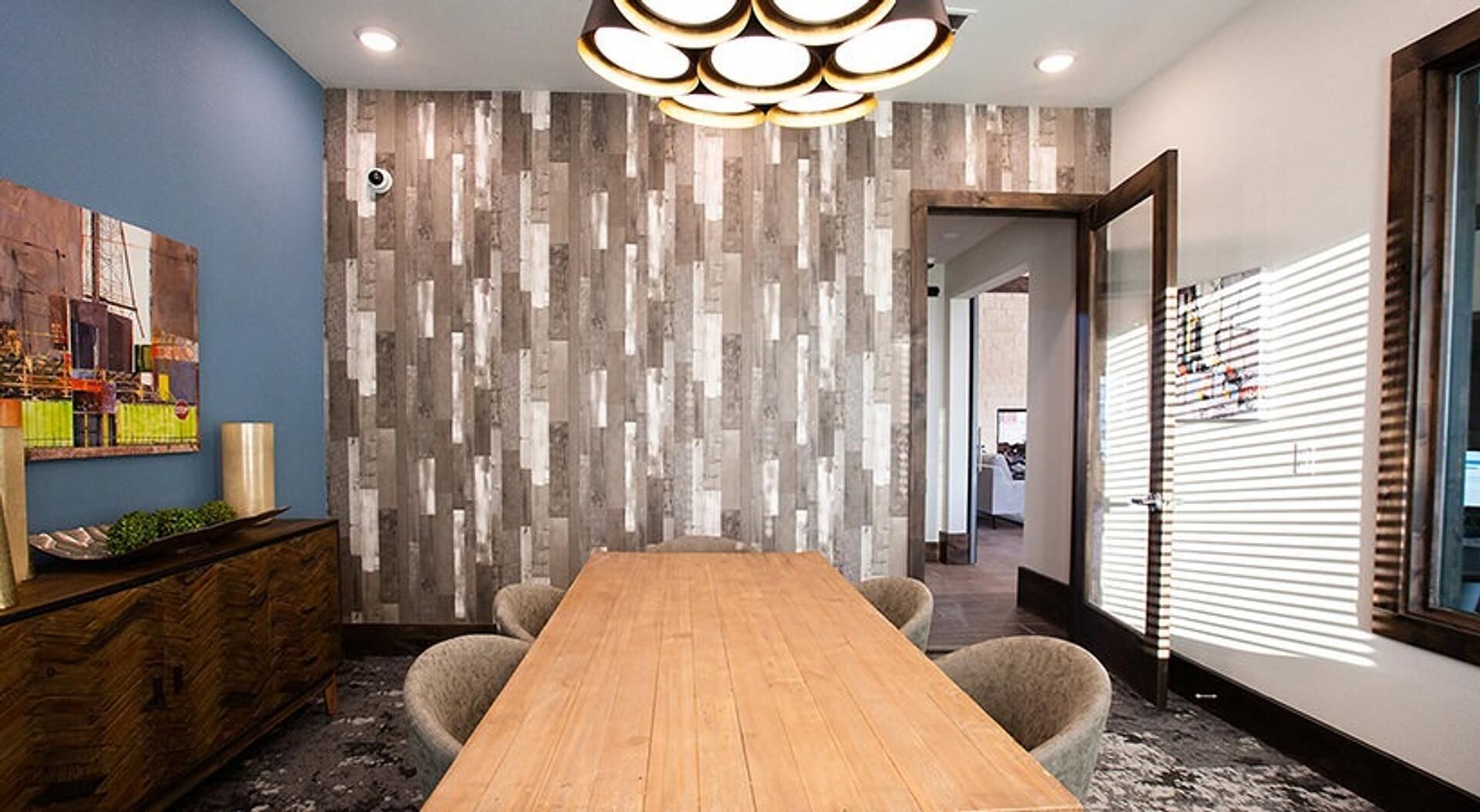 Uma sala de conferência com mesa de madeira, cadeiras, luminária moderna, parede decorativa e janela com persianas.