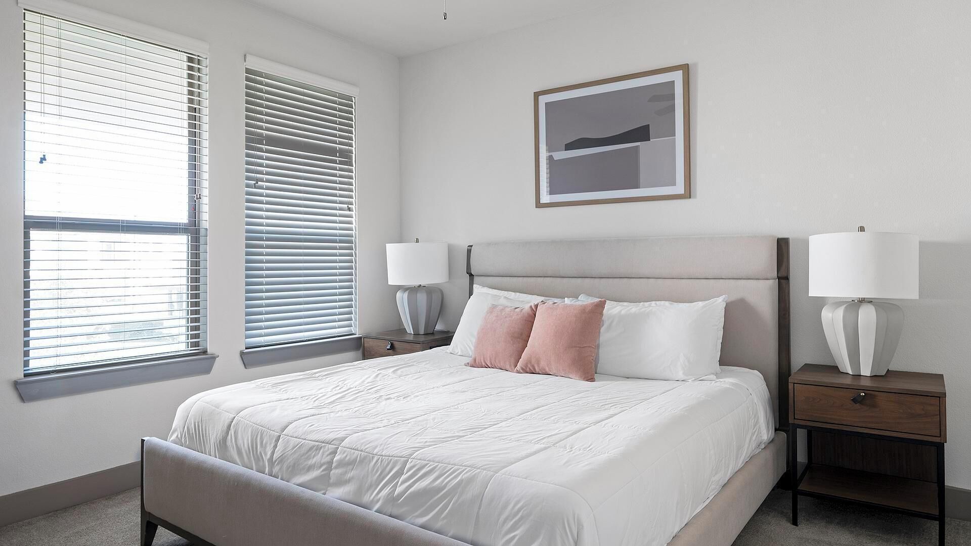 A bedroom with a bed, two bedside tables with lamps, a window with blinds, and a framed picture on the wall.