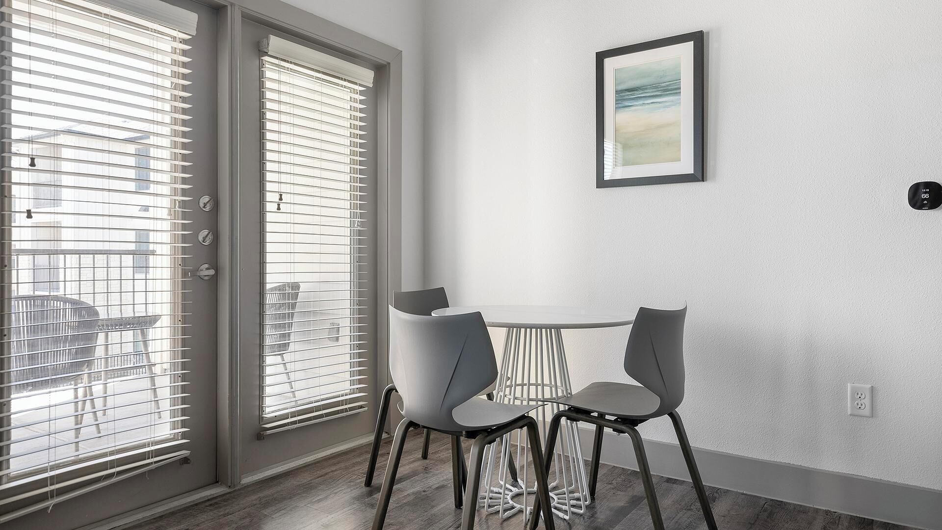 A modern room with a sliding glass door, a small round table with two chairs, a framed picture on the wall, and a thermostat.