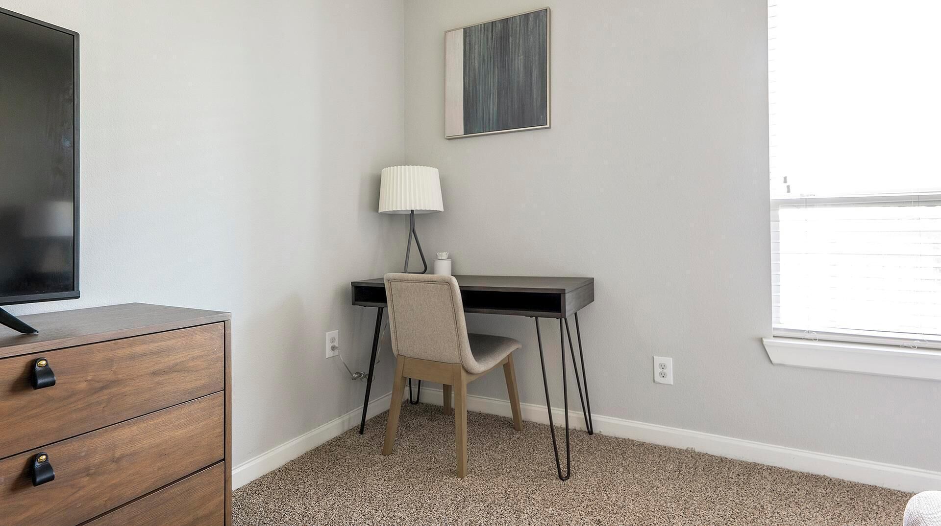 Una habitación con un escritorio de madera, una silla, un televisor, una lámpara y una ventana.
