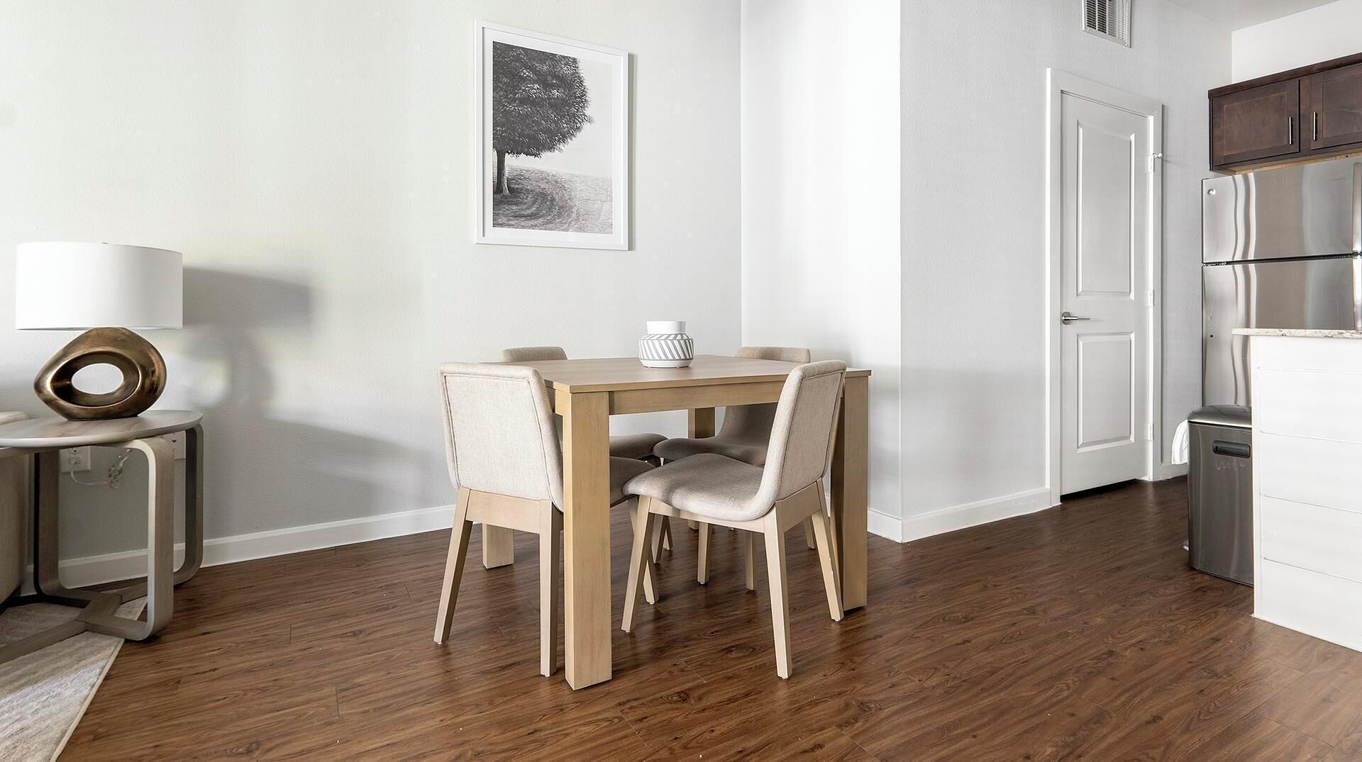 A dining area with a wooden table and chairs, a small side table with a lamp, a framed picture on the wall, and a kitchen area with a refrigerator and cabinets.